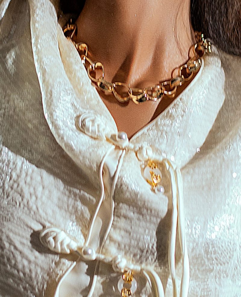 Close detail of knotted fabric closures with pearl buttons and a heavy gold chain at the neckline.