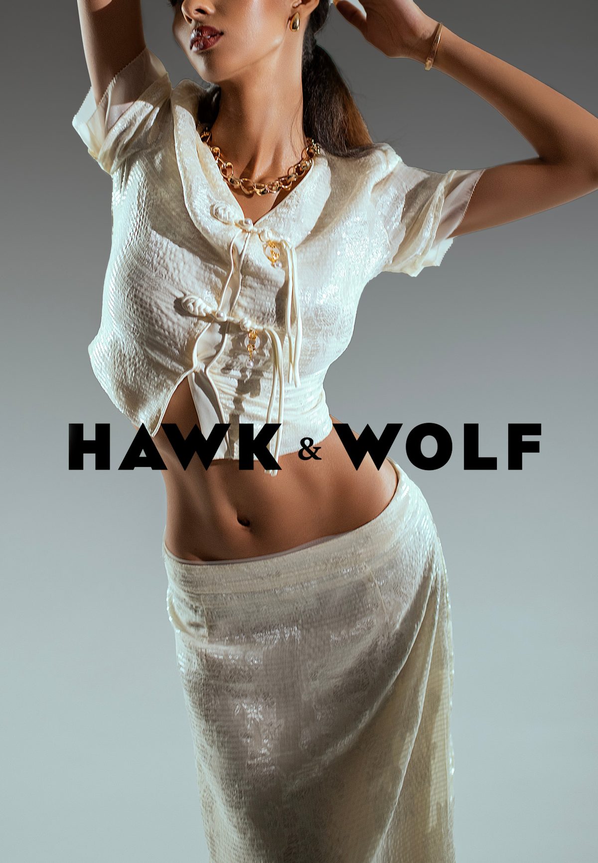 The Hawk & Wolf wordmark over a model in an ivory sequinned top with knotted closures, a matching skirt and a gold chain.