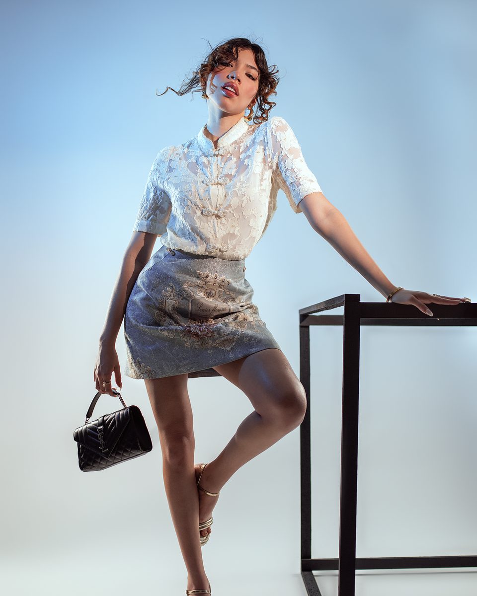 A model in an ivory jacquard shirt with knotted closures and a grey floral mini skirt, holding a black quilted bag beside a black steel frame.